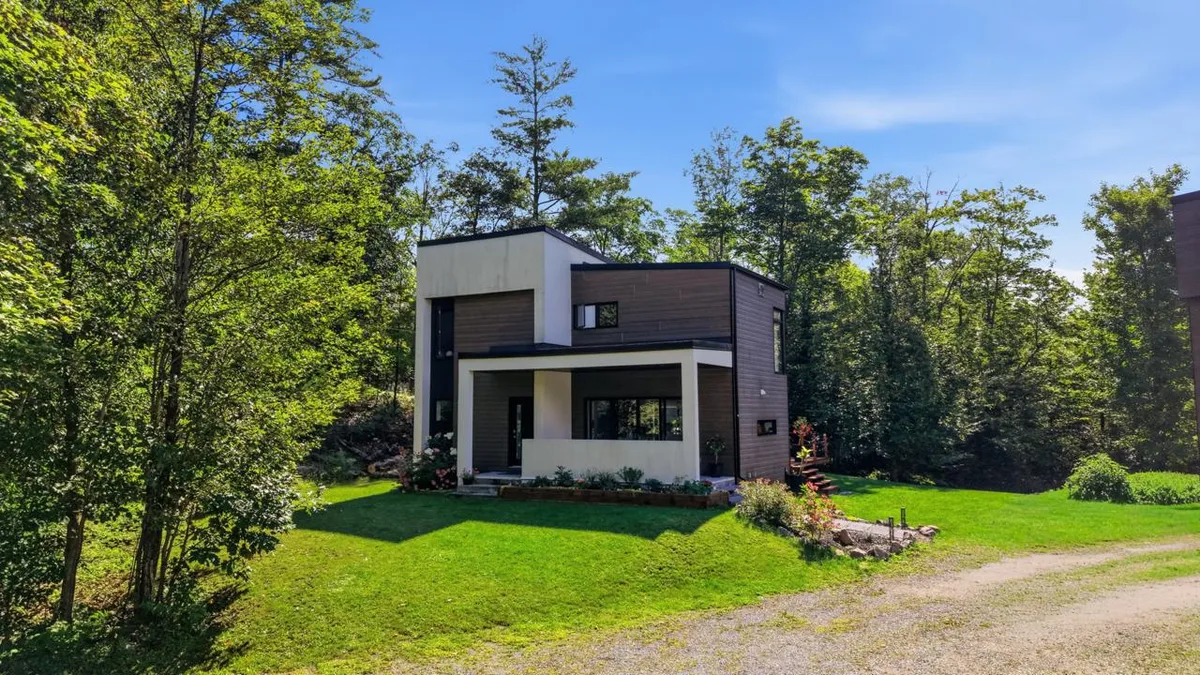 Contemporary two-story house in a wooded clearing with dark siding, white-framed porch, and green lawn